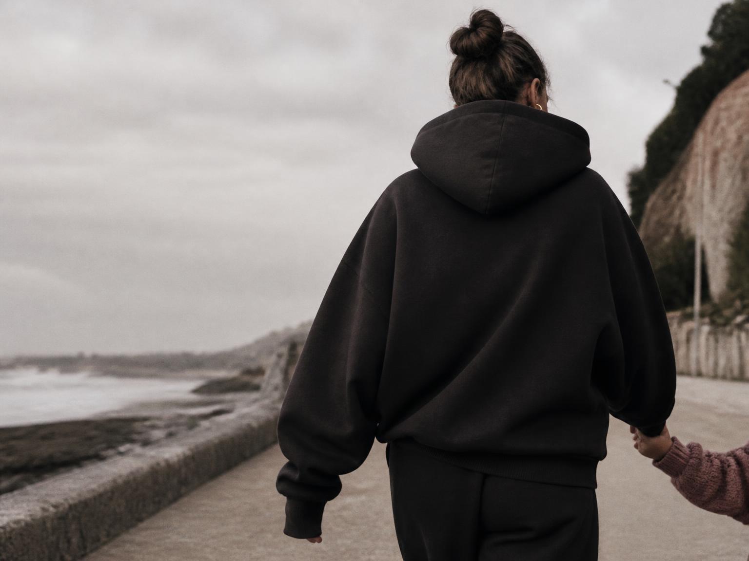 A mother in an oversized black hoodie walking beside the coast, holding her child’s hand