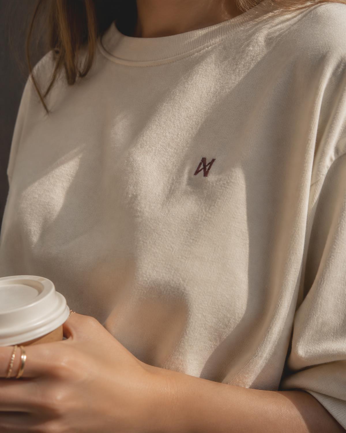 A subtle burgundy mark embroidered over the heart of a bone cotton tee
